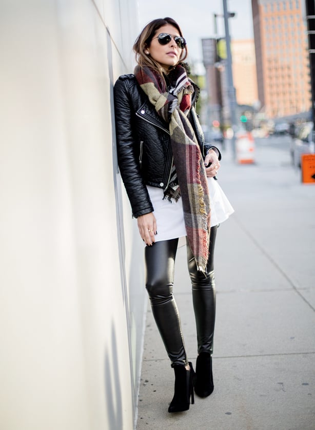 A woman leans against a wall, wearing a white mini skirt and full-length leather leggings with stiletto boots 