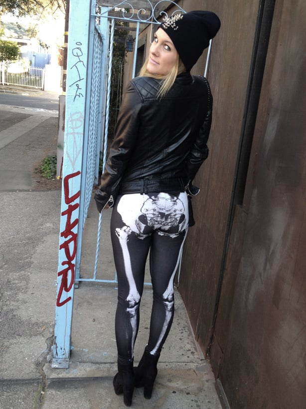 A blonde model wearing black skeleton print leggings, a leather jacket and a black woolly hat