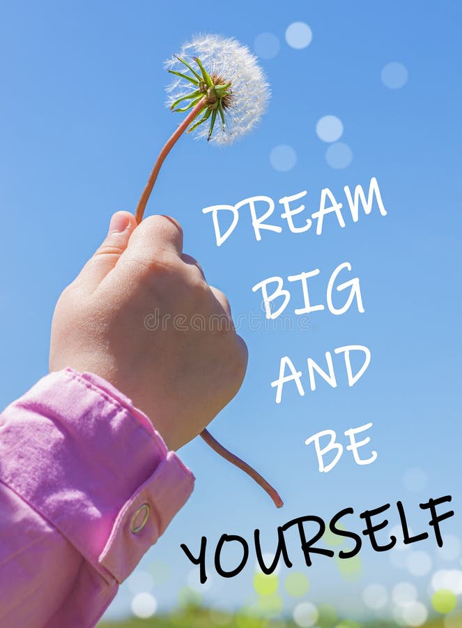 Dream Big and Be Yourself text sign. White fluffy dandelion flower in the hand of a little child on blue sky background royalty free stock photography