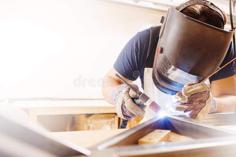 Male in face mask welds with argon-arc welding. Male in face mask, protective gloves welds with argon-arc welding. Welder makes weld seam on metal frame. Worker royalty free stock photos