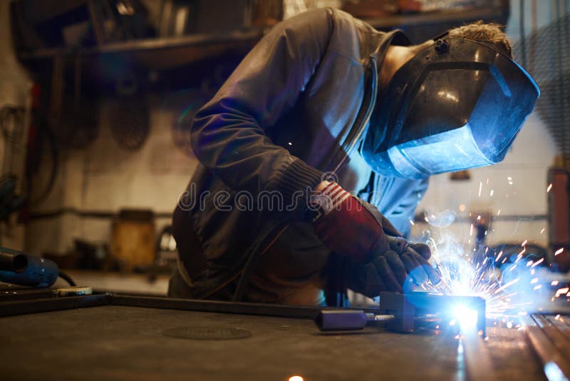 Welding iron workpieces. Young welder in protective mask leaning over workbench and welding iron workpieces royalty free stock image